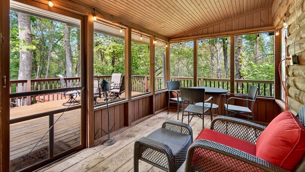 A covered outdoor deck with wooden flooring, metal chairs, a table, and a sofa with red cushions.