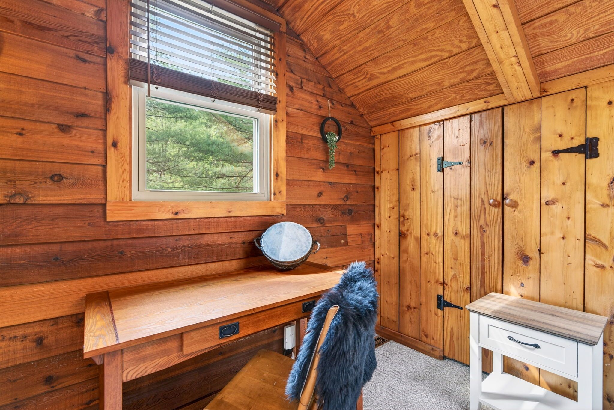 L’intérieur d’un chalet en bois, avec une fenêtre, un banc en bois, une chaise recouverte d’un plaid en fourrure et une petite table blanche.