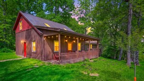 A wooden cabin with a red door, a porch with string lights, and a fenced yard.