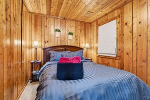 A wooden-paneled room with a bed, two bedside tables with lamps, a window with blinds, and two wall-mounted planters.