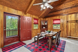 Un intérieur en bois comprenant une table à manger, des chaises, un ventilateur de plafond et une vue sur l’extérieur, visible à travers une porte.