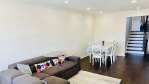 Un salon avec un canapé d’angle gris, une table à manger blanche avec des chaises et un escalier menant à l’étage supérieur.