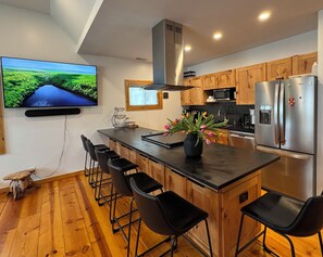 A modern kitchen with wooden cabinets, a stainless steel refrigerator, a flat-screen TV, and a bar area with stools. - Tulip Tree House in Shorewood-Tower Hills-Harbert, Michigan (Shorewood-Tower Hills-Harbert)