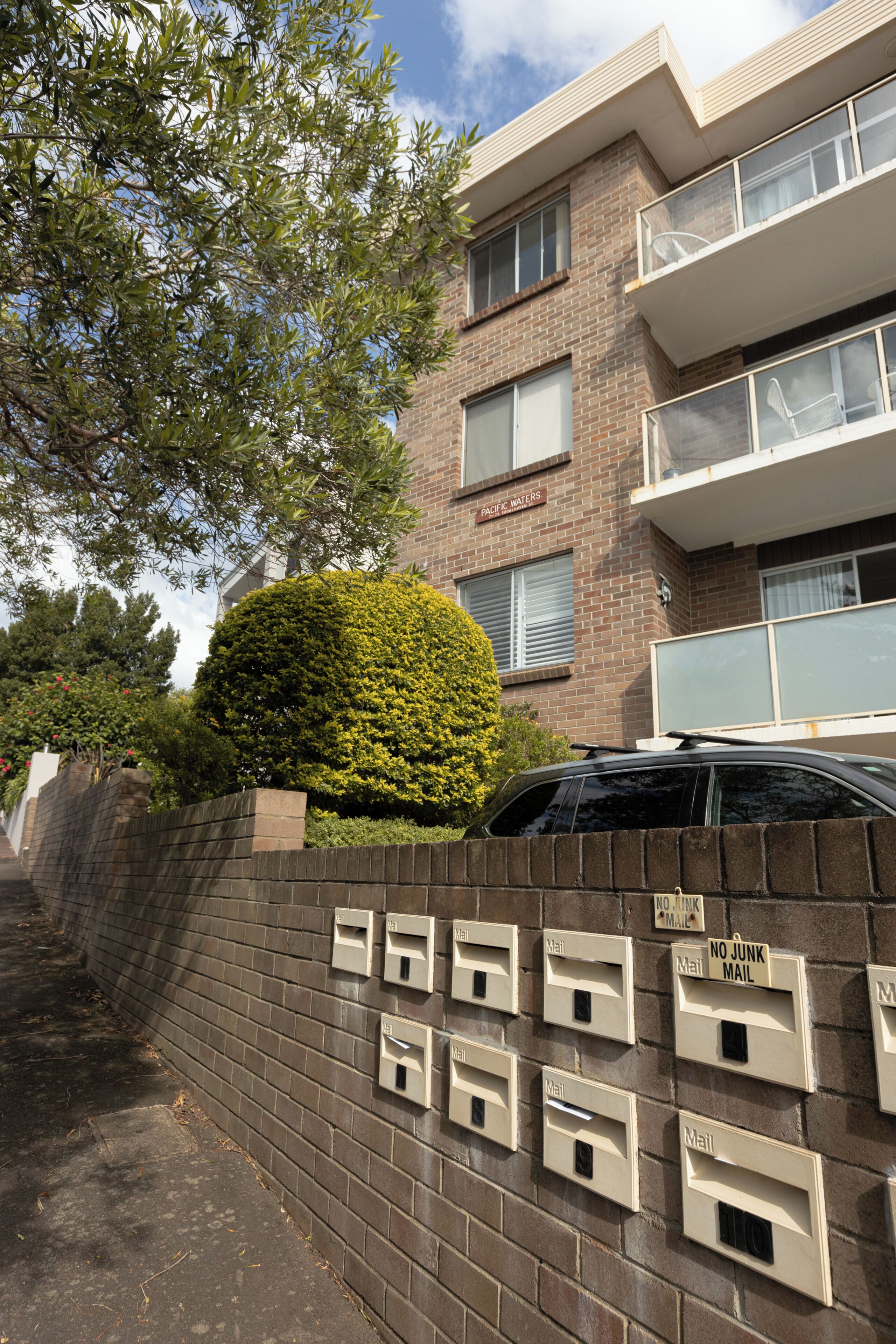 A multi-story building with mail slots on the wall, a hedge, and a car parked in front.