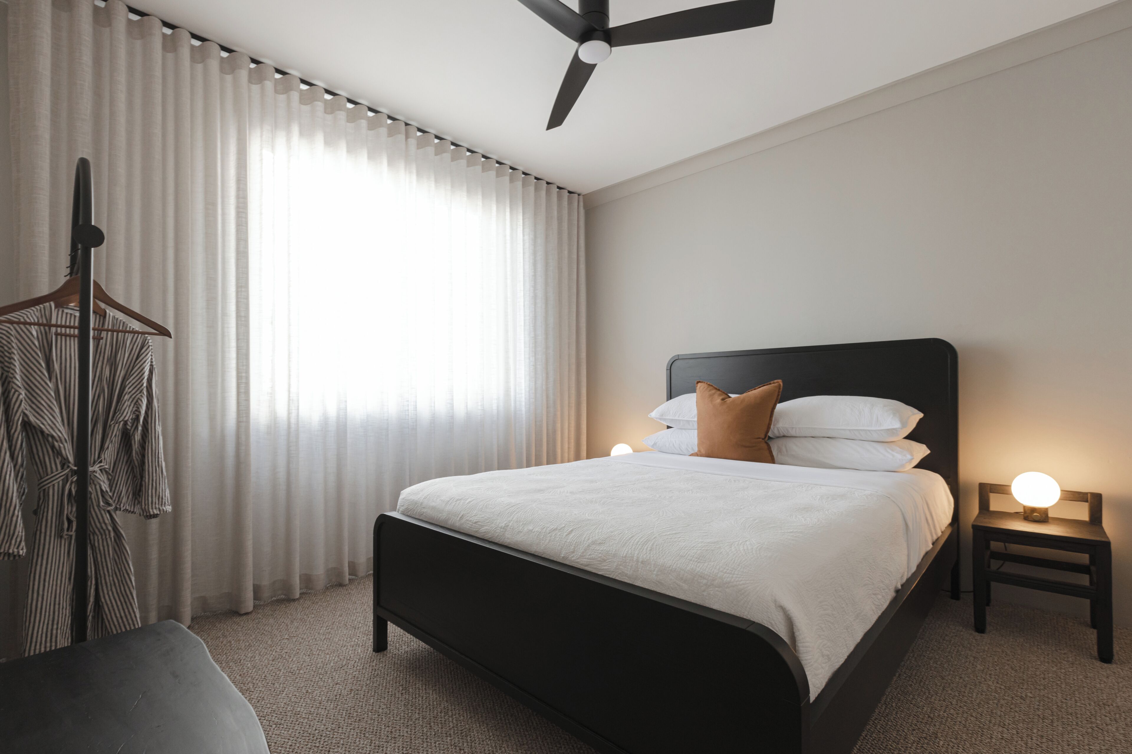 A hotel room with a bed, a bedside table with a lamp, a hanging coat rack, and a window with curtains.