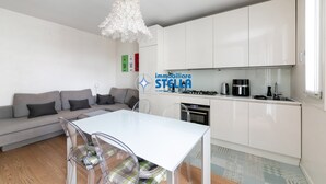 A modern living room with a sofa, a dining table with chairs, a kitchen area with cabinets and appliances, and a decorative white chandelier. - Maricel residence °swimming pool#modern° (Lido di Jesolo)