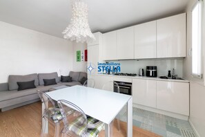 A modern living room with a sofa, a dining table with chairs, a kitchen area with cabinets and appliances, and a decorative white chandelier. - Maricel residence °swimming pool#modern° (Lido di Jesolo)