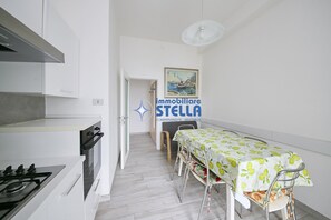 Una cucina con mobili bianchi, un piano cottura e un'area pranzo con tavolo e sedie. Sul muro si nota un cartello e un quadro sopra l'area pranzo.