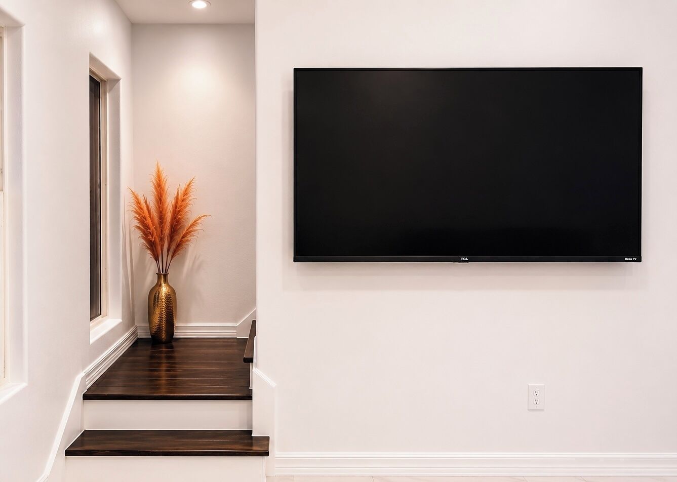 Una escalera con escalones de madera oscura y un jarrón con plumas de pampa anaranjadas. Un televisor de pantalla plana está montado en la pared sobre la escalera.