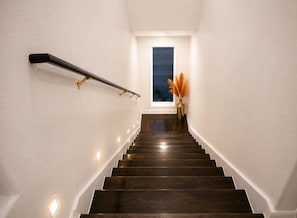 Uma escadaria com degraus de madeira, corrimão, luminárias na parede e um vaso com plumas de capim-do-campo.