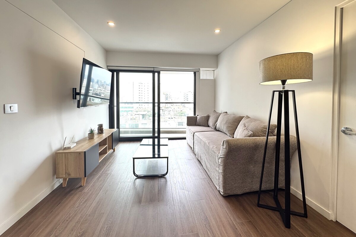 A modern living room with a sofa, a standing lamp, a TV mounted on the wall, and a window with a city view.