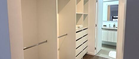 A walk-in closet with open shelving and drawers, a wooden floor, and a view into a bathroom with a sink and mirror.