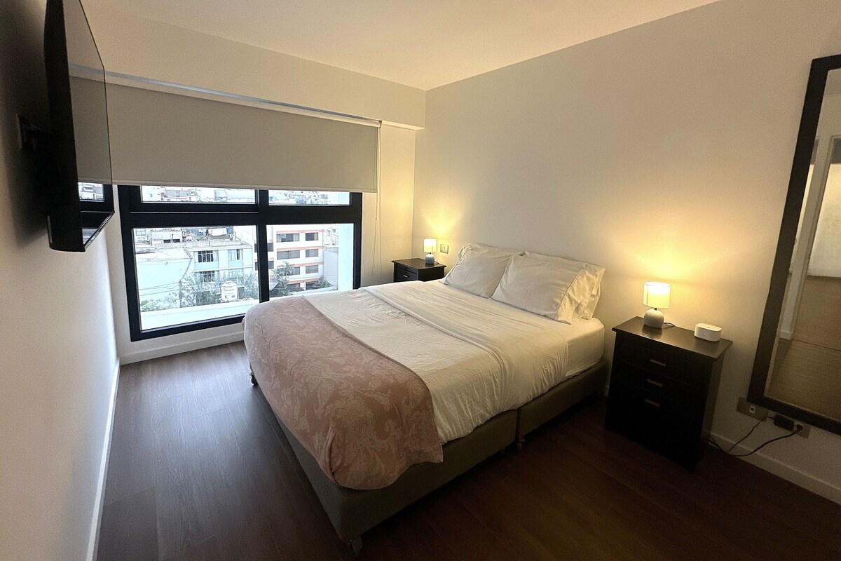 A bedroom with a bed, bedside tables, a window, and a flat-screen TV mounted on the wall.