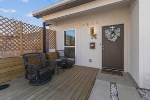 Une terrasse en bois avec des chaises en osier et une petite table, une porte portant le numéro 2811 et une clôture en treillis.