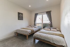 Une chambre avec deux lits, une fenêtre avec des rideaux, une citation inspirante sur le mur et un sol en béton.