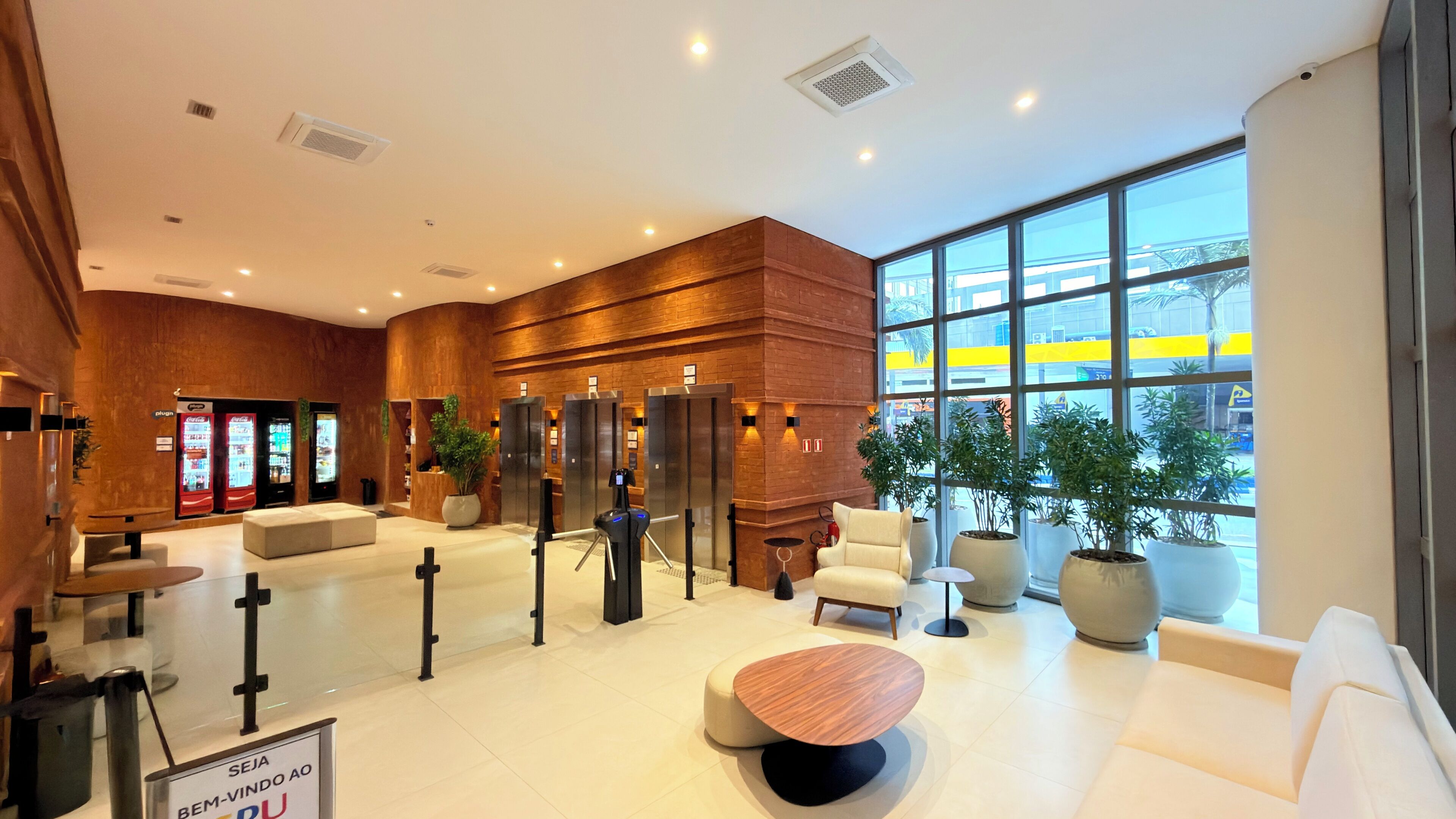 A modern lobby with a reception desk, seating area, vending machines, elevators, and large windows offering a view of the outside.