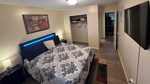 Une chambre à coucher comprenant un lit, une table de chevet, une lampe, un tableau, une armoire et un téléviseur.