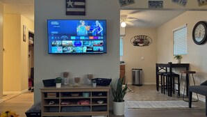 Un salon doté d’un téléviseur à écran plat fixé au mur, d’un meuble de rangement en bois, d’un coin repas avec une table et des chaises, et d’une cuisine située à l’arrière-plan.