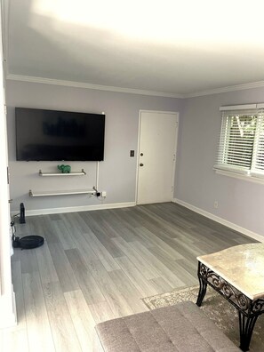 Ein Zimmer mit einem an der Wand montierten Flachbildfernseher, einer Tür, einem Fenster mit Jalousien, einer kleinen Ablage und einem Tisch mit gemustertem Tischläufer.