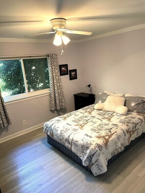 Ein Schlafzimmer mit Bett, Deckenventilator, Fenster mit Vorhängen und Nachttisch mit Lampe.