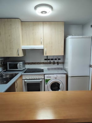 A compact kitchen with wooden cabinets, a microwave, stove, oven, washing machine, and a refrigerator. - Nice apartment with garage in Conil. (Conil De La Frontera)