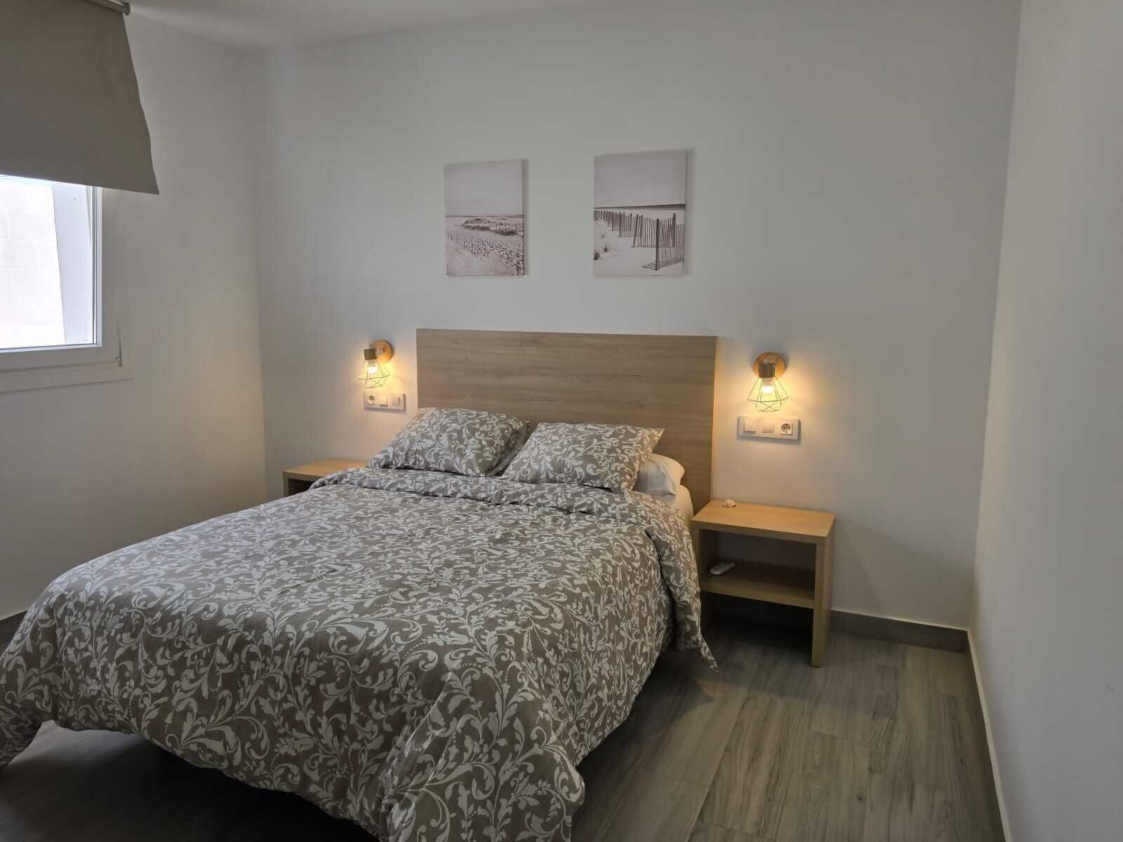 Un dormitori amb un llit, dues taules de nit, làmpades penjades a la paret i dos quadres emmarcats.