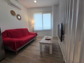 Ein Wohnzimmer mit einem roten Sofa, einem kleinen weißen Tisch, einem an der Wand montierten Fernseher und einem Fenster mit Jalousien.