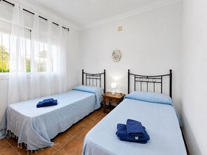 Ein Schlafzimmer mit zwei Einzelbetten, einem Nachttisch mit Lampe und einem Fenster mit durchsichtigen Vorhängen.