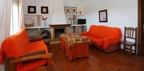 Ein Wohnzimmer mit zwei orangefarbenen Sofas, einem Holzstuhl, einem kleinen Tisch mit buntem Tischläufer, einem Kamin und einem Fernseher auf einem Schrank.