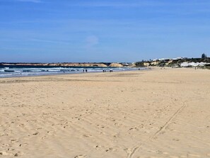 Ein weiter Sandstrand mit Fußspuren, die zum Wasser führen, ein klarer Himmel und Gebäude im Hintergrund.