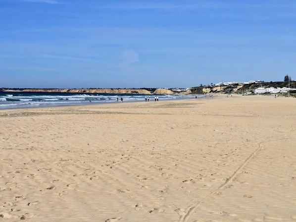Ein weiter Sandstrand mit Fußspuren, die zum Wasser führen, ein klarer Himmel und Gebäude im Hintergrund.
