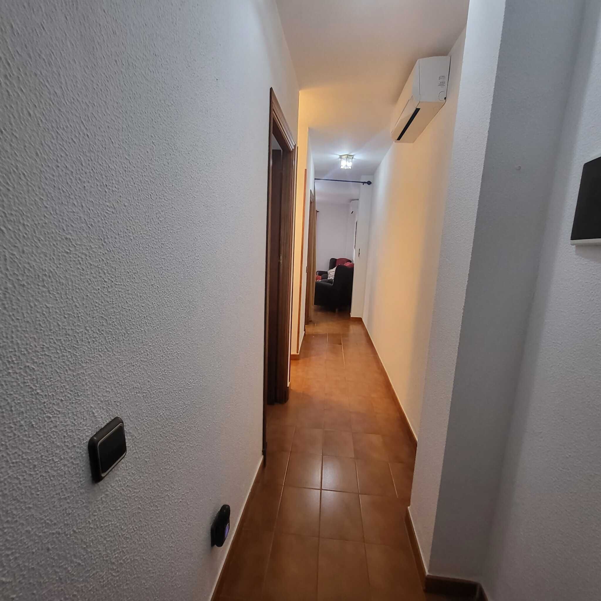 Een smalle gang met witte wanden, een houten vloer en een deur aan het einde die leidt naar een kamer met een bed en een stoel.
