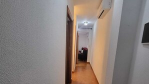 Een smalle gang met witte wanden, een houten vloer en een deur aan het einde die leidt naar een kamer met een bed en een stoel.