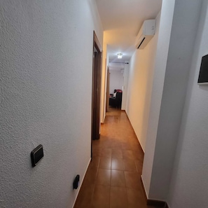 Ein schmaler Korridor mit weißen Wänden, Holzboden und einer Tür am Ende, die in einen Raum mit Bett und Sessel führt.
