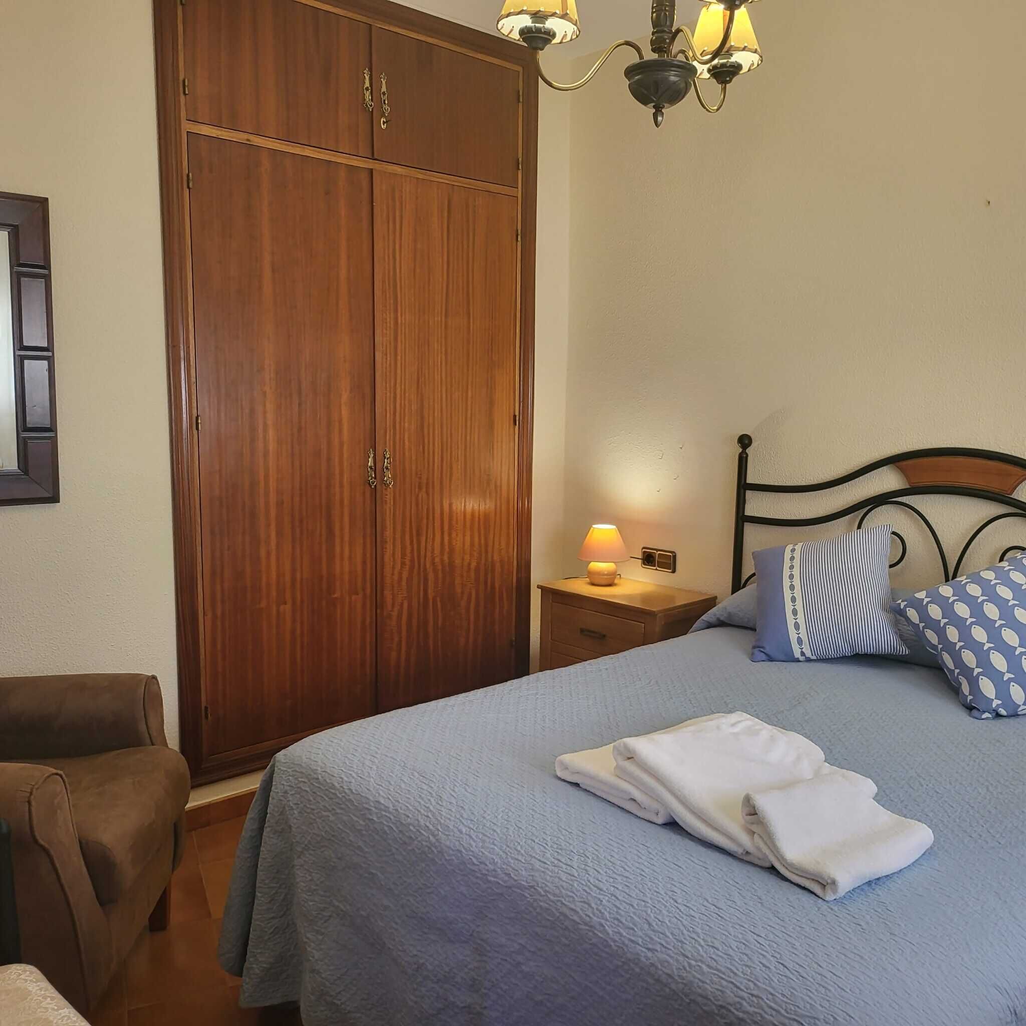 Een slaapkamer met een bed, kussens, een nachtkastje, een lamp, een kast en een stoel.