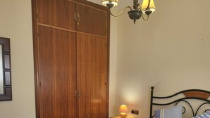 Een slaapkamer met een bed, kussens, een nachtkastje, een lamp, een kast en een stoel.