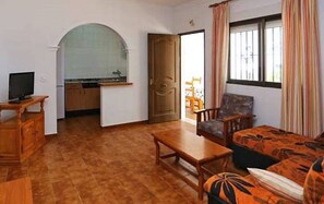 Ein Zimmer mit einer Couch, einem Holztisch, einem Fernseher und einer Küchenzeile mit Kühlschrank, sowie einer Tür zu einem weiteren Raum.