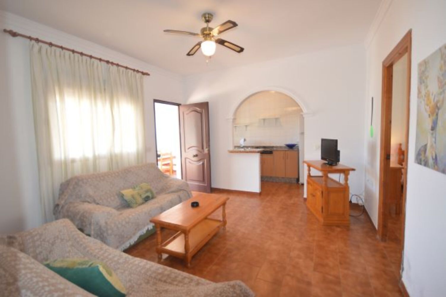 Un salon comprenant un canapé, une table basse, un téléviseur posé sur un meuble en bois, un ventilateur de plafond et un coin cuisine visible à travers une arche.