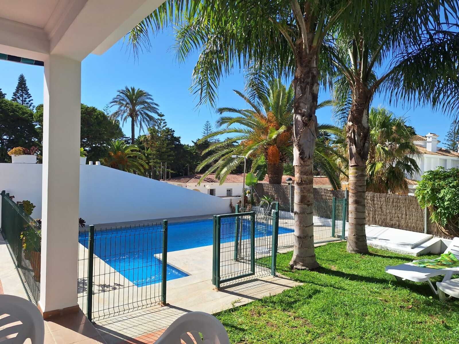 Un área de piscina con piscina vallada, palmeras y un edificio blanco al fondo.