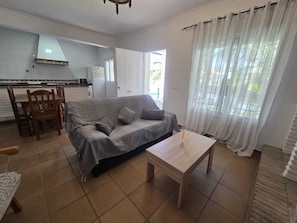 A living room with a sofa, a coffee table, a dining table with chairs, a kitchen area, and a large window with curtains. - Coquettish house with private area near the sea (Conil De La Frontera)