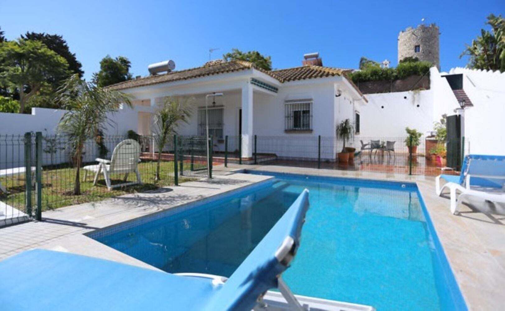 A swimming pool with a clear blue water, a white building with a tiled roof, and a tower in the background.