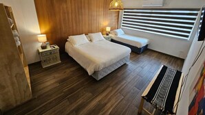 A bedroom with a large bed, a smaller bed, a nightstand, a lamp, a wooden wall, a window with blinds, and a hanging light fixture. - Hotel Casa Ellies Boutique (Puerto Varas)