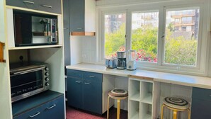 A compact kitchen with a microwave, toaster, and coffee maker on the counter, a window with a view of greenery, and a small stool. - Hotel Casa Ellies Boutique (Puerto Varas)