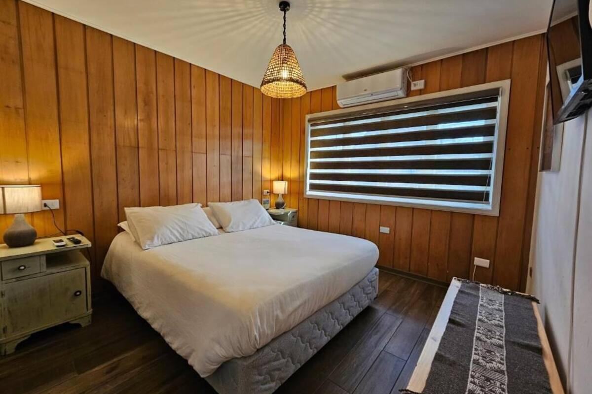 Un dormitorio con cama, paredes de madera, una ventana con persianas, una lámpara colgante y un equipo de aire acondicionado.