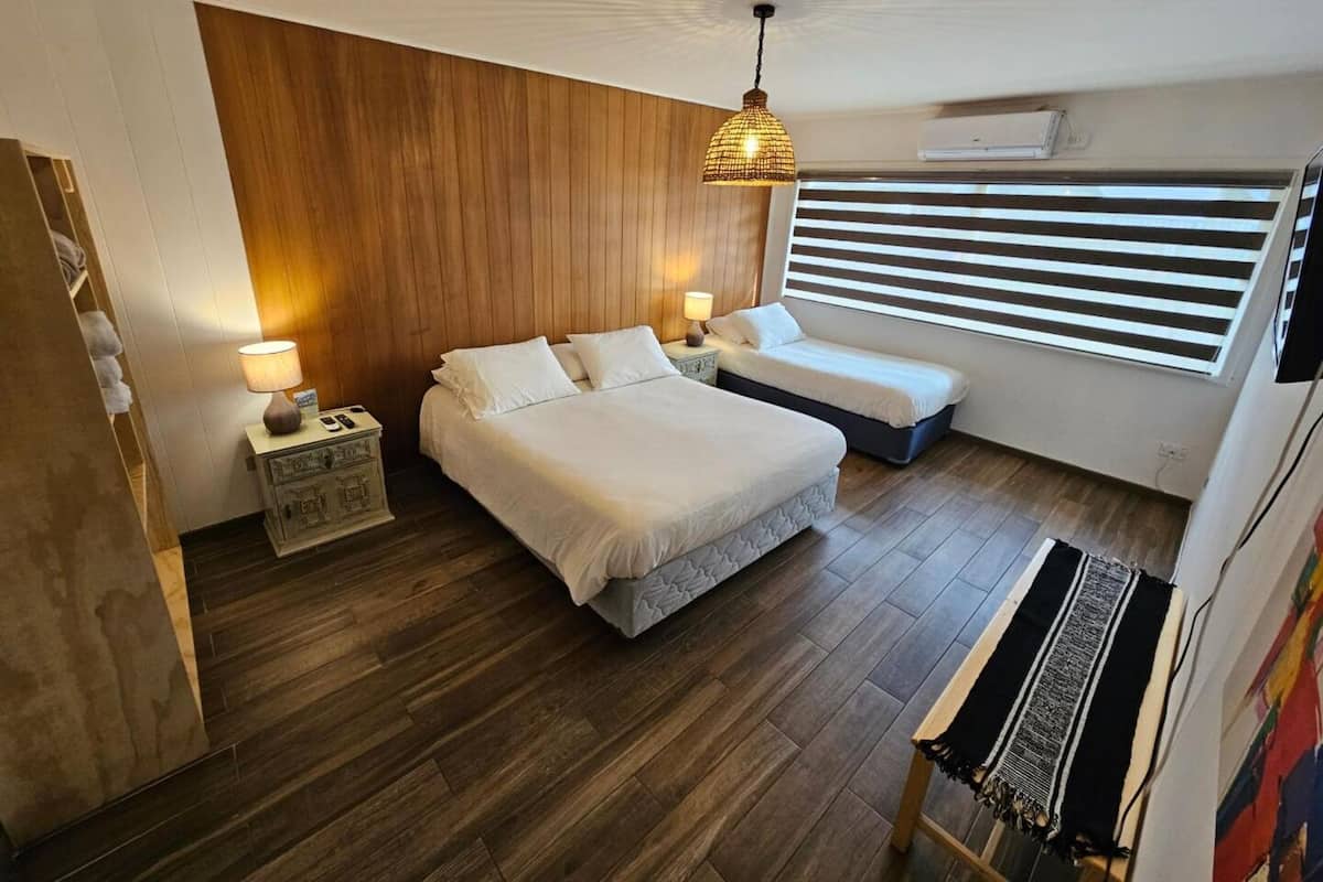 A bedroom with two beds, a wooden wall, a hanging light fixture, a window with blinds, and a bench.