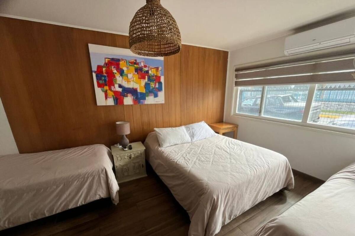 Habitación de hotel con dos camas, cabecera de madera, cuadro abstracto de colores, lámpara de fibra tejida y ventana con vista a coches estacionados.