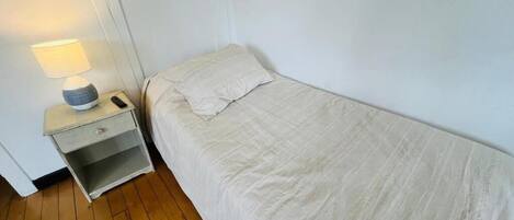 Una cama individual con edredón blanco, una mesita de noche con lámpara y un mueble auxiliar con cajón.