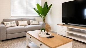Uma sala de estar com um sofá cinzento, uma mesa de centro, uma planta grande e uma televisão em um suporte.