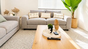 Uma sala de estar com um sofá cinzento, uma mesa de centro, uma planta grande e uma janela com persianas.