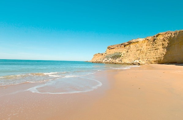 Ein Strand mit klarem, blauem Wasser, sandigem Ufer und einer felsigen Klippe.